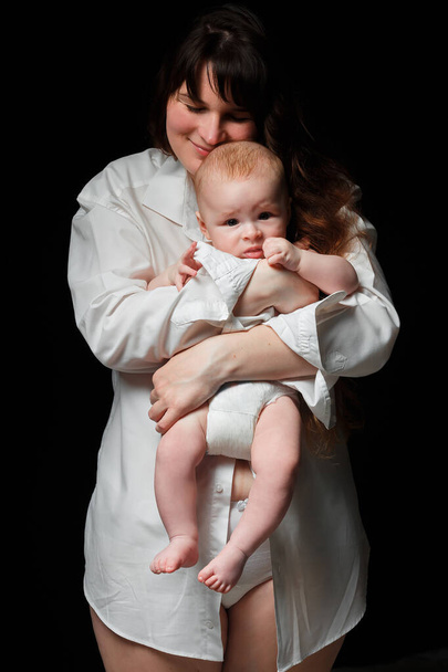 giovane mamma bruna con i capelli lunghi e una camicia bianca tiene un figlio appena nato. Un bambino di 4 mesi tra le braccia. Su uno sfondo nero
. - Foto, immagini