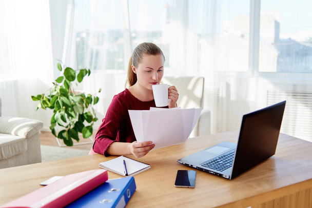 Retrato de una joven mujer de negocios confiada sentada en el escritorio de madera, bebiendo café y leyendo documentos en la oficina en casa, espacio para copiar
 - Foto, imagen