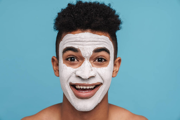 Photo of excited african american man in facial cosmetic mask smiling at camera isolated over blue background - Foto, imagen