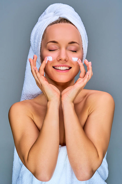 Happy woman in a white bath towel with a white towel on her hair with cream on cheeks - Photo, image