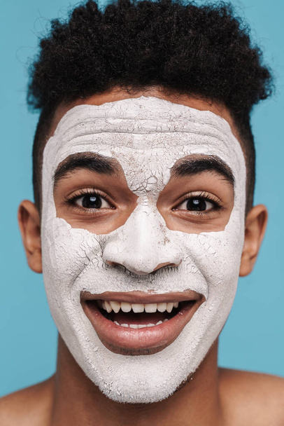 Foto de hombre afroamericano excitado en máscara cosmética facial sonriendo a la cámara aislada sobre fondo azul
 - Foto, imagen