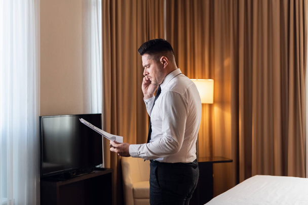 Rano w luksusowym hotelu. Widok z boku przystojnego młodego biznesmena wykonującego telefon, stojącego w pewnej pozie przy panoramicznym oknie z jakimiś planami w ręku - Zdjęcie, obraz
