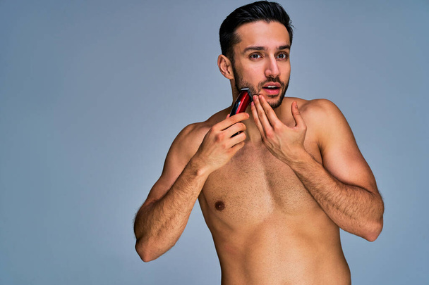 Naked man with black hair with a beard with red electric shaver in hand - Zdjęcie, obraz