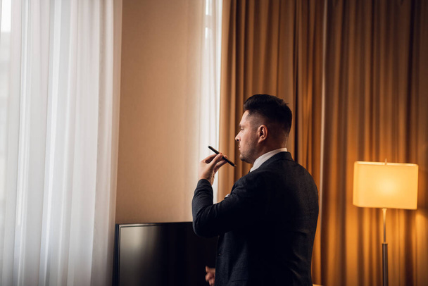 Seitenansicht eines gut aussehenden jungen Geschäftsmannes, der in selbstbewusster Pose vor einem Panoramafenster seines Hotelzimmers steht und telefoniert - Foto, Bild