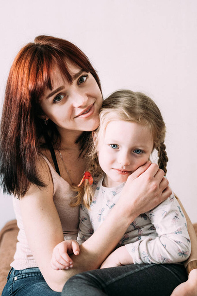 Rozmazaný portrét matky a dcery na bílém pozadí. Máma brunetka drží svou blond dceru v náručí, zatímco sedí na gauči. Matčina péče o dítě. Koncept dne matek. - Fotografie, Obrázek