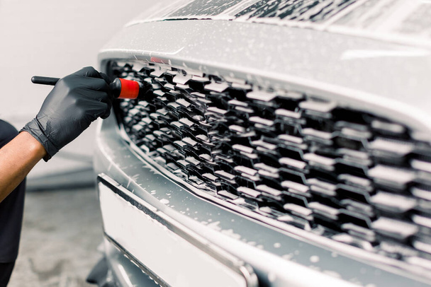 Car detailing wash. Detailing cleaning with soap at car wash. Cropped image of hand of male worker in rubber gloves washing car radiator grille of luxury white car with special brush - Foto, Imagem