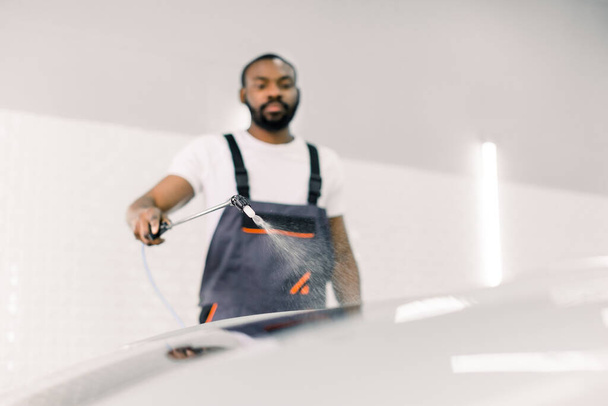 Auto service, car washing and cleaning concept. Handsome young Afrian American male worker in t-shirt and gray overalls, doing car prewash with high pressured water gun. Selective focus on cleaner - Foto, Bild