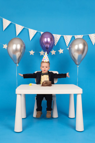 Happy caucasian boy is sitting at table in festive cone-shaped hat at birthday party, rejoicing and raising his hands to side, celebrating victory and achieving goal, on decorated blue background. - Zdjęcie, obraz