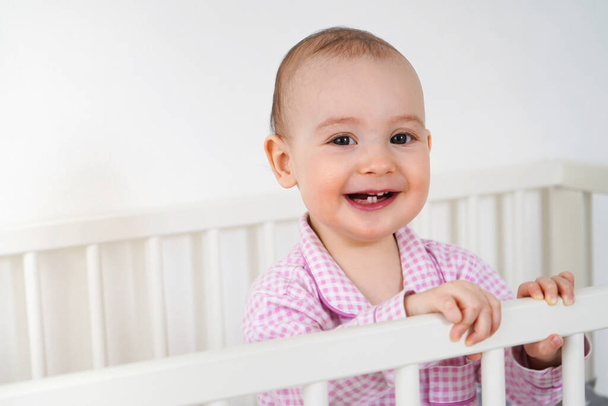 Joyful baby in pink pajamas is standing in the crib. The child stood on its own legs and smiles. Baby is glad to see her mom - Valokuva, kuva