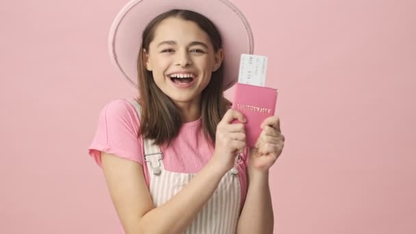 Happy pretty brunette woman in overalls and hat holding passport with tickets and looking at the camera over pink background - Video