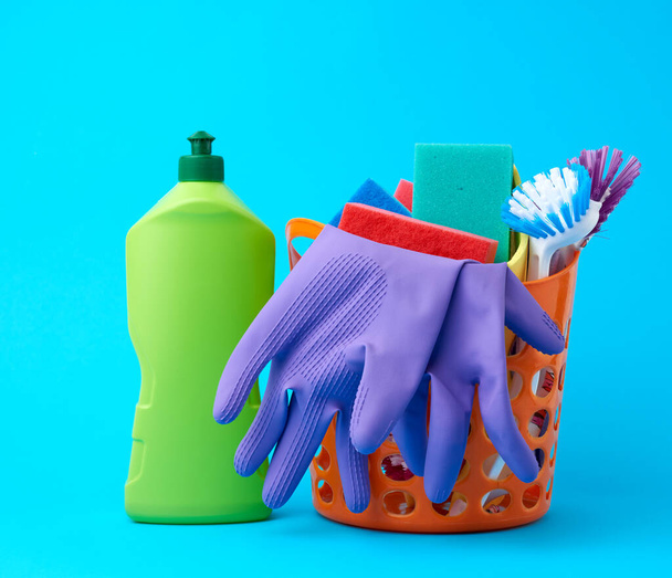 оранжевая корзина со стиральными губками, резиновыми защитными перчатками, щетками и чистящим средством в зеленой пластиковой бутылке на синем фоне, комплект
 - Фото, изображение