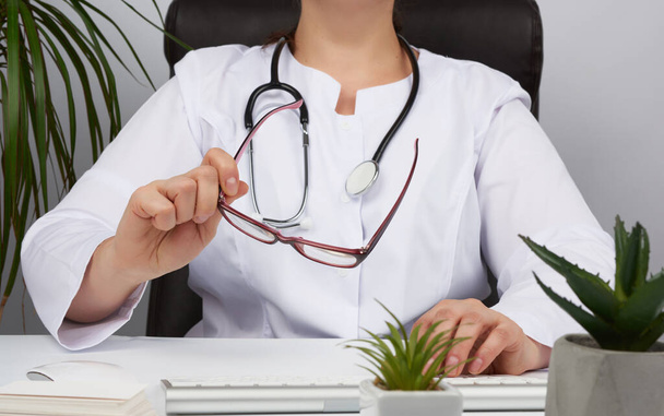 Ärztin in weißer Uniform, medizinische Einmalmaske sitzt auf einem Stuhl am Tisch und arbeitet, hält eine Brille in der Hand, Büro - Foto, Bild
