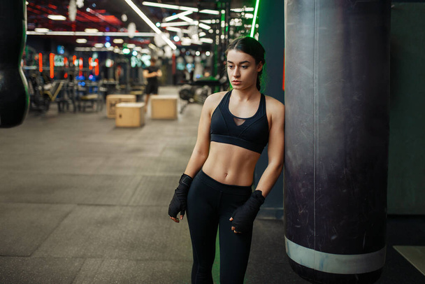 Γυναίκα με μαύρους επιδέσμους ποζάρει στο σάκο του μποξ, εκπαιδεύεται σε κουτιά. Γυναίκα πυγμάχος στο γυμναστήριο, κορίτσι kickboxer στο αθλητικό σύλλογο, kickboxing προπόνηση - Φωτογραφία, εικόνα