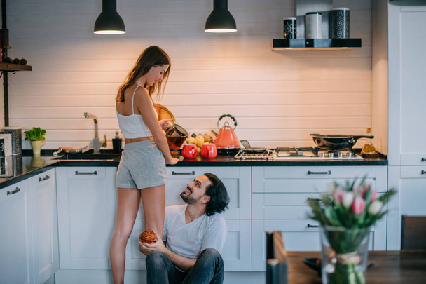 Νεαρό ζευγάρι το πρωί στην κουζίνα στο σπίτι. Άνδρας και γυναίκα έχουν ένα διασκεδαστικό πρωινό σε μια όμορφη κομψή κουζίνα. - Φωτογραφία, εικόνα