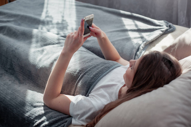 Young and beautiful woman laying on bed in morning light and using smartphone to connect to internet and use social media with happy and relaxed manner. Cheerful woman holding mobile phone. - Fotó, kép
