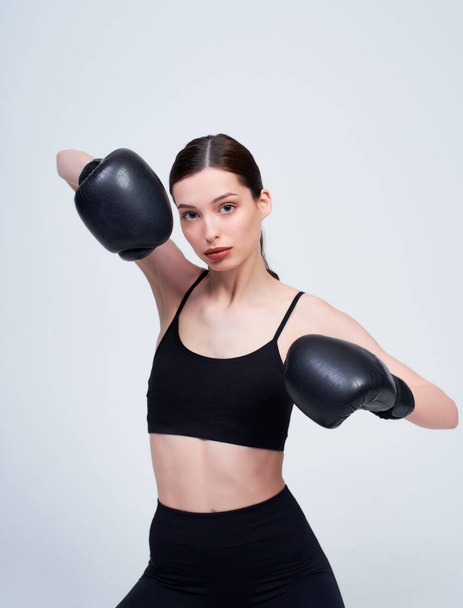 Photo of a healthy 20s fitness woman in black sportswear posing on camera in black boxing gloves while training in the gym, isolated on white background. Sport concept, healthy lifestyle, dream body. - Photo, image