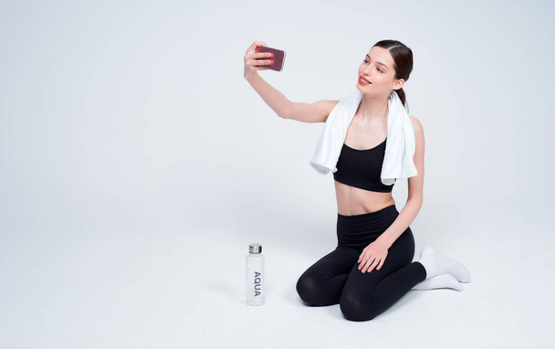 Attractive happy young Caucasian woman doing yoga, fitness, pilates, gymnastics, in a black sports suit, sits in white space, resting after exercise, takes a selfie. - Photo, image