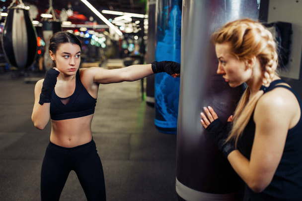 Nainen nyrkkeilysiteissä lyö nyrkkeilysäkkiä ja treenaa valmentajan kanssa. Naisnyrkkeilijät kuntosalilla, potkunyrkkeilyharjoitus urheiluseurassa, lyöntiharjoitukset
 - Valokuva, kuva
