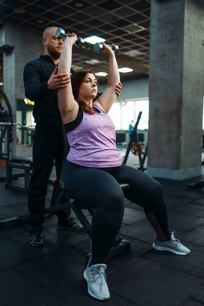 Nadwaga kobieta z trenerem robi ćwiczenia z hantlami w klubie sportowym, trening fitness z instruktorem. Kobieta zmaga się z nadwagą, trening tlenowy przeciwko otyłości, siłownia - Zdjęcie, obraz