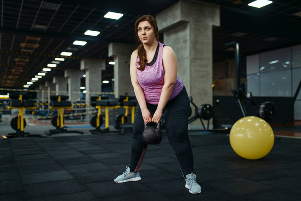 Kilolu bir kadın spor salonunda kettlebell ile egzersiz yapıyor, aktif antrenman yapıyor. Obez kadın aşırı kilolarla, obeziteye karşı aerobik egzersizlerle, spor kulüpleriyle mücadele eder. - Fotoğraf, Görsel