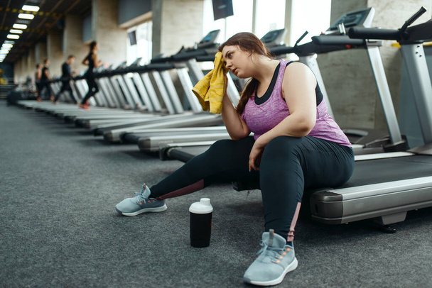 Zmęczona kobieta z nadwagą siedząca na bieżni na siłowni, wypoczynek po aktywnym treningu. Otyła kobieta zmaga się z nadwagą, trening tlenowy przeciwko otyłości, klub sportowy - Zdjęcie, obraz