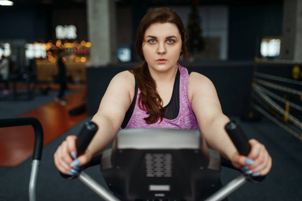 Υπέρβαρη γυναίκα προπονείται με ποδήλατο γυμναστικής, ενεργή προπόνηση. παχύσαρκη γυναίκα παλεύει με το υπερβολικό βάρος, αερόβια προπόνηση κατά της παχυσαρκίας, αθλητικός σύλλογος - Φωτογραφία, εικόνα