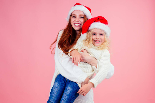 ,happy redhead ginger woman and cute little blonde girl wearing santa claus hat and having fun together in studio pink background - Photo, Image