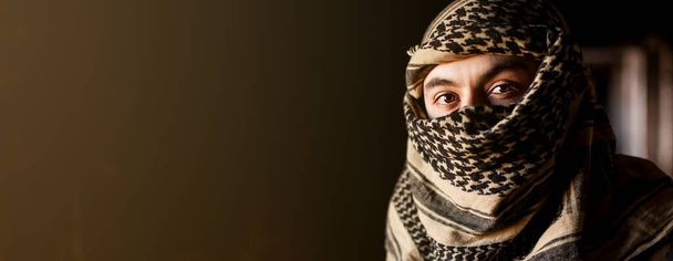 Portrait of arab man in keffiyeh on face. Muslim with a sad face in a military jacket and a headdress from the national keffiyeh on a black background. - Photo, image