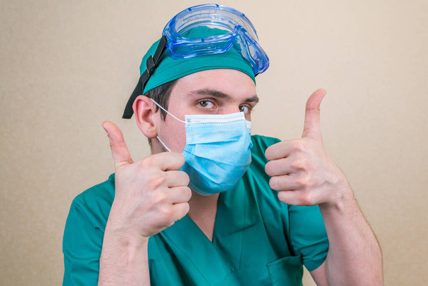 Dokter met duimen omhoog in groen pak en blauwe masker handschoenen en beschermende bril op gele achtergrond. Selectieve focus. - Foto, afbeelding
