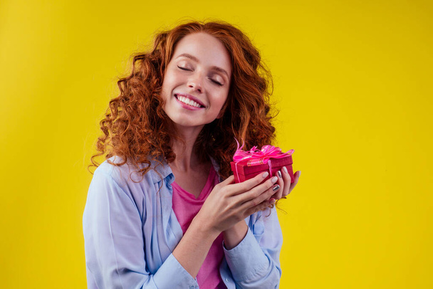 redhair curly ginger woman holding gift box in studio yellow background.mothers day and eighth of marchor St. Valentines Day concept - Фото, зображення