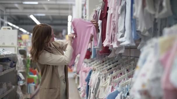 szczęśliwa młoda kobieta wybrać ubrania dla dzieci, piękna kobieta w sklepie próbuje jakości odzieży, sklep z artykułami dla dzieci - Materiał filmowy, wideo