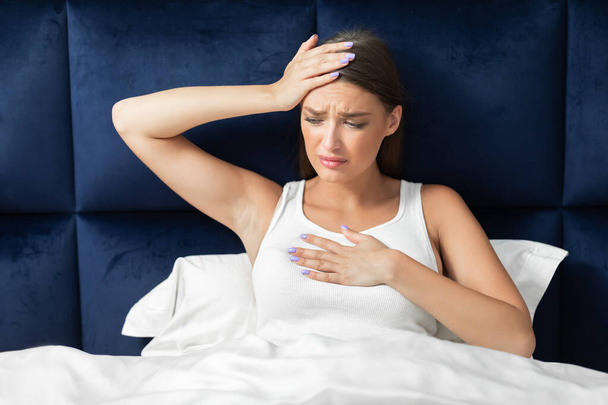 Женщина с лихорадкой и затрудненным дыханием сидит в постели в помещении
 - Фото, изображение