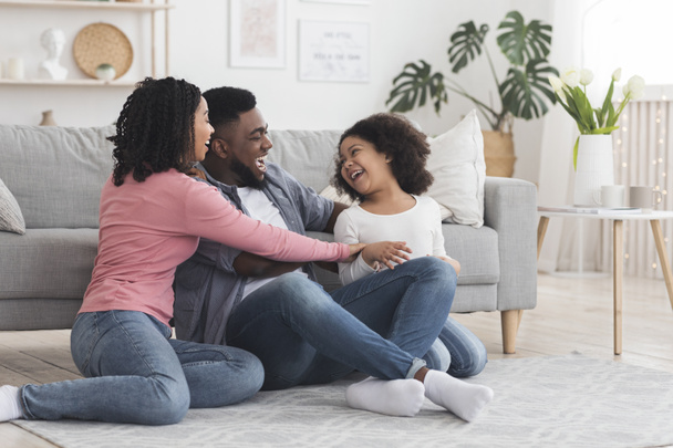 Cheerful black family having fun at home, parents tickling their daughter - Fotó, kép