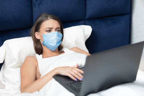 Chica trabajando en portátil usando máscara protectora acostado en la cama
 - Foto, Imagen