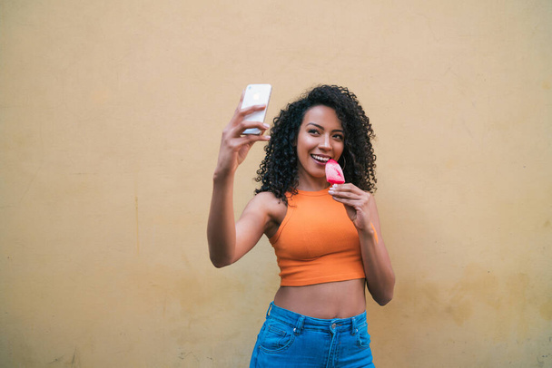 Portrait of afro woman taking selfies with her mophile phone while eating ice-cream. Technology and lifestyle concept. - Foto, Imagem