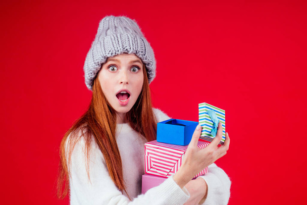redhaired ginger woman wear stylish wool hat and white sweater looking at camera and holding three boxes witg gifts copyspase in studio red background - Photo, image