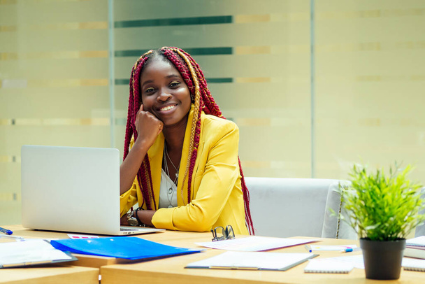 happy, afro american woman in a stylish yellow jacket and multi-colored dreadlocks pigtails workplace in a light large modern office - Фото, зображення