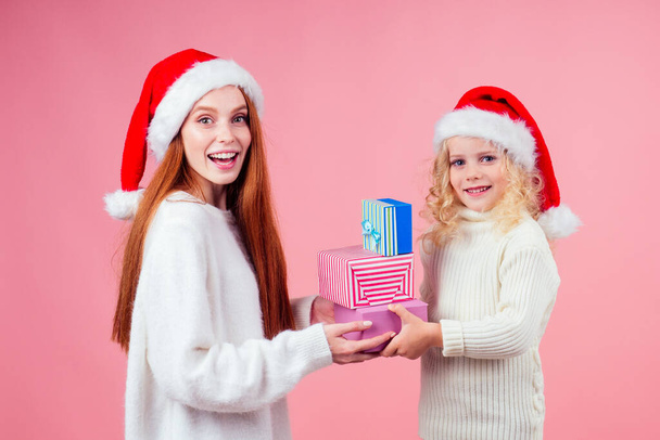 getting xmas presents.girls in red santa hat holding a bunch of gifts boxes in studio pink background - Фото, изображение