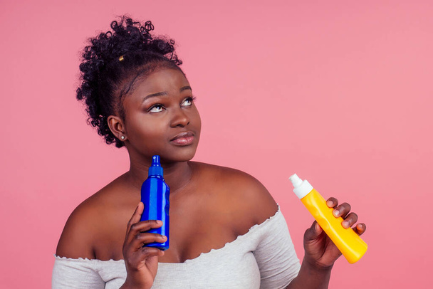 african american woman applying cream shoosing between two products in studio pink background - Fotó, kép