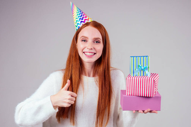 redhaired ginger woman wearing birthday cap horn holding many gift boxes in studio white background - Fotoğraf, Görsel