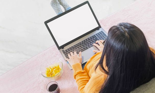Jonge mooie Aziatische vrouw droeg oranje gebreide kleding, zitten op een grijze bank, werken online levensstijl, ontspannen thuis met koffiemok, thuis werkende laptop, werk van thuis uit - Foto, afbeelding