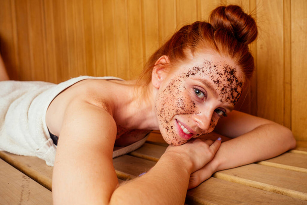 redhaired ginger young woman peeling face with brown sugar in traditional russian banya.steaming and self-care in finnish sauna - Fotoğraf, Görsel