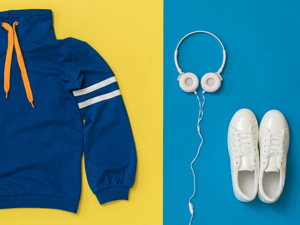 Witte hoofdtelefoon, witte sneakers en een blauw sportjasje op een gele en blauwe achtergrond. - Foto, afbeelding