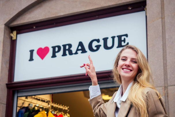 Beautiful girl in red dress travel in Praha. She finger pointing at storefront with text i love prague - Photo, Image