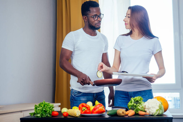 Korean woman wearimg white cotton t-shirt with her african american boyfriend cooking in fry pan bresksast - Foto, Imagen