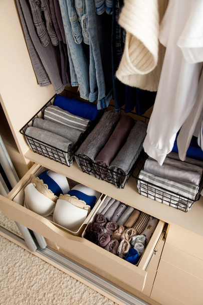 Top view of neatly folded things and accessories in boxes and drawers lying under things hanging on hangers in a large wardrobe. Concept of accuracy and general cleaning in the house - Фото, зображення