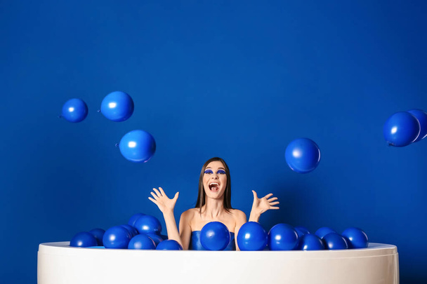 Portret van modieuze jonge vrouw met luchtballonnen zittend in bad op blauwe achtergrond - Foto, afbeelding