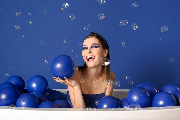 Portrait of fashionable young woman with air balloons and soap bubbles sitting in bathtub on blue background - Фото, зображення