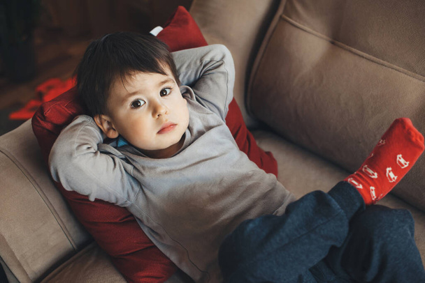 Górny widok portret białego chłopca odpoczywającego na kanapie i spoglądającego w górę do kamery podczas kwarantanny - Zdjęcie, obraz
