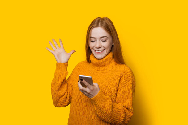 Fehér vörös lány szeplős gesztikulál a győzelem gazdaság egy mobil és pózol egy meleg pulóver sárga alapon - Fotó, kép